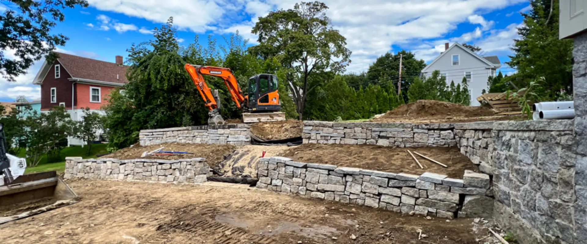 An orange excavator from Hines Ground Services working on landscaping with tiered stone retaining walls in a residential area with houses and trees.