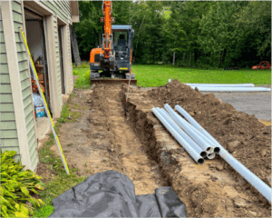 An excavator digging a trench next to a house with PVC pipes laid nearby, ready for installation by Hines Ground Services.