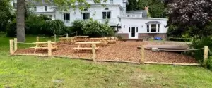 A fenced outdoor area with wood chip flooring, featuring several wooden picnic tables and a round sandbox, set against a backdrop of a white house and trees.