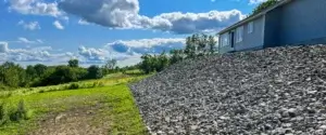 A rural landscape featuring a house on a gravel hill with a lush green field under excavation by an excavating contractor, set against a blue sky with fluffy clouds.